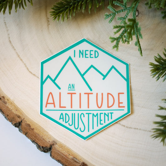 Hexagon shaped sticker with text "I need an Altitude Adjustment." Blue and pink elements on a cream colored background