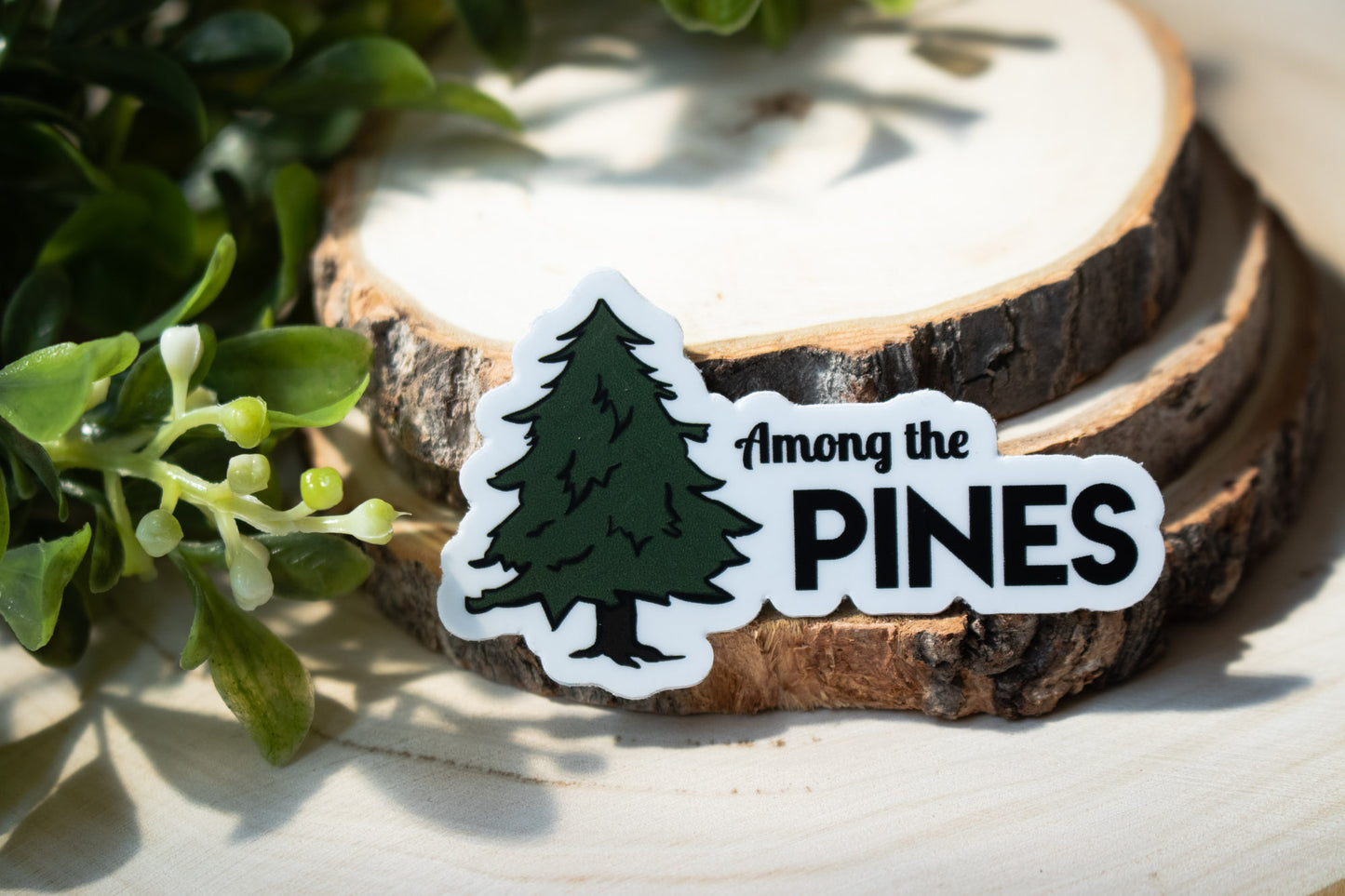 White sticker with forest green pinetree illustration and txt reading "among the pines"