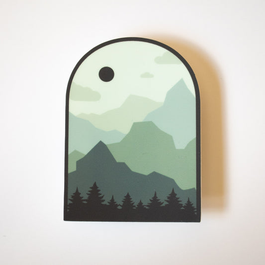 Arch shaped sticker in various shades of green depicting a layered mountain scene
