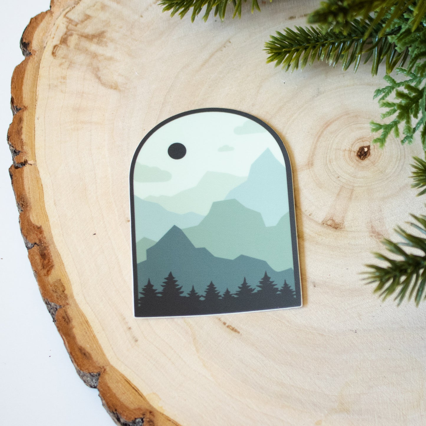 Rounded top sticker showing pine trees, layers of mountain, and a full moon